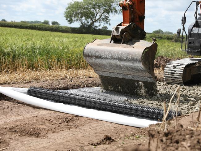 A digger pouring aggregate onto a newly laid roll of geogrid